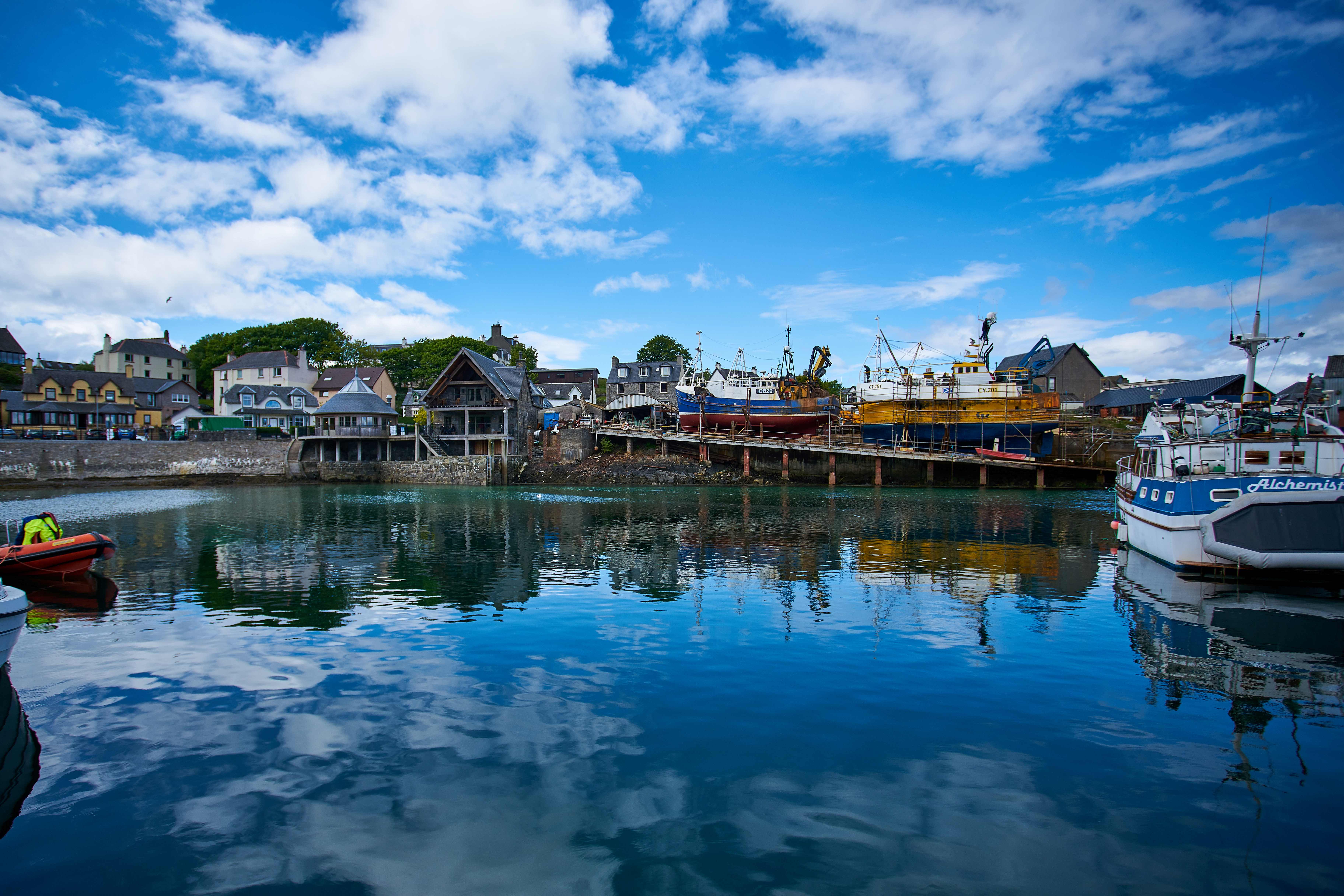 A picturesque scene of Mallaig Harbour with boats, set beneath a bright blue sky filled with white clouds.