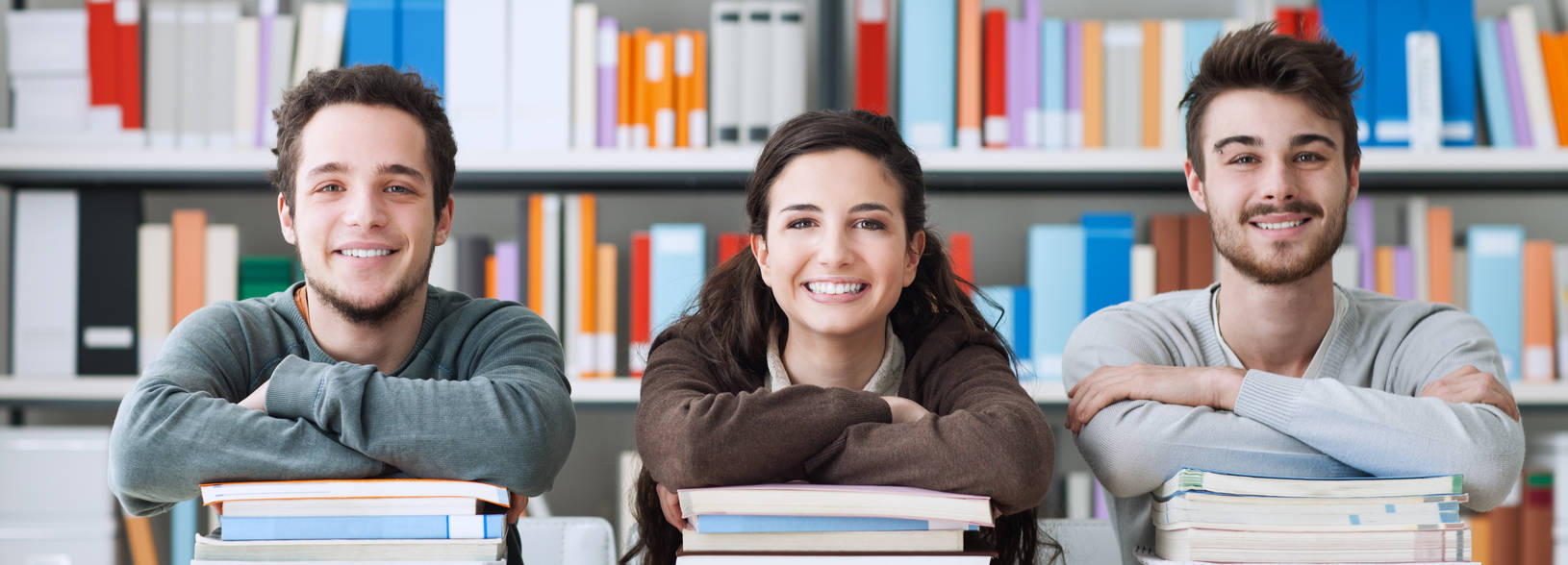 Three students resting their elbows on piles of books 